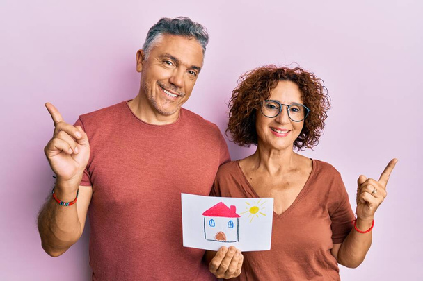 Красивая пара среднего возраста, держащая дома рисовать покупая дом для будущего улыбаясь счастливый указывая рукой и пальцем в сторону  - Фото, изображение