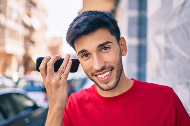 Giovane uomo latino sorridente felice ascolto messaggio audio utilizzando smartphone in città. - Foto, immagini