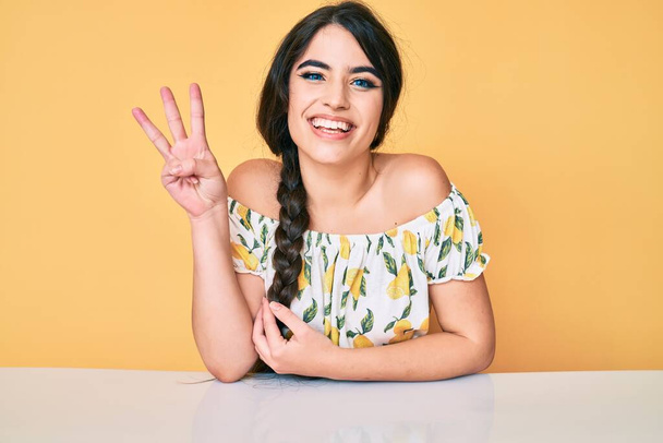 Ragazza bruna adolescente che indossa abiti casual seduta sul tavolo mostrando e indicando con le dita il numero tre mentre sorride fiducioso e felice.  - Foto, immagini
