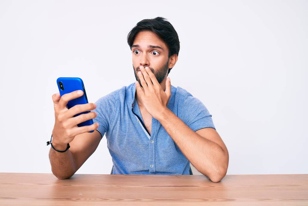 Knappe Latijns-Amerikaanse man met behulp van smartphone zitten op de tafel bedekken mond met de hand, geschokt en bang voor fouten. verraste uitdrukking  - Foto, afbeelding