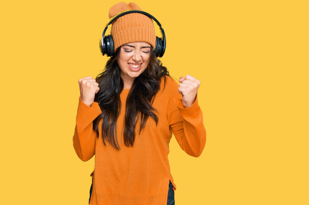 Gyönyörű barna fiatal nő hallgat zenét a fülhallgató izgatott siker karokkal és csukott szemmel ünnepli győzelem mosolyogva. győztes koncepció.  - Fotó, kép