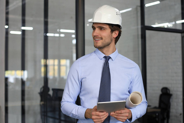 Confident young business man in hardhat holding blueprint and looking away while standing indoors - Photo, Image