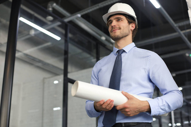 Confident young business man in hardhat holding blueprint and looking away while standing indoors - Photo, Image