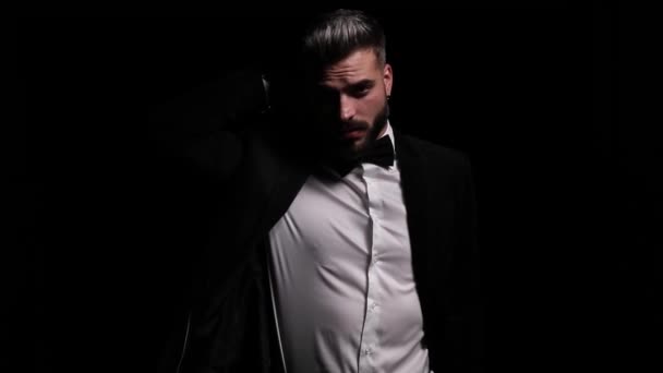elegant young man adjusting black tuxedo, holding hand behind neck and posing, moving in a side view position and looking over shoulder and disappearing in the dark - Footage, Video