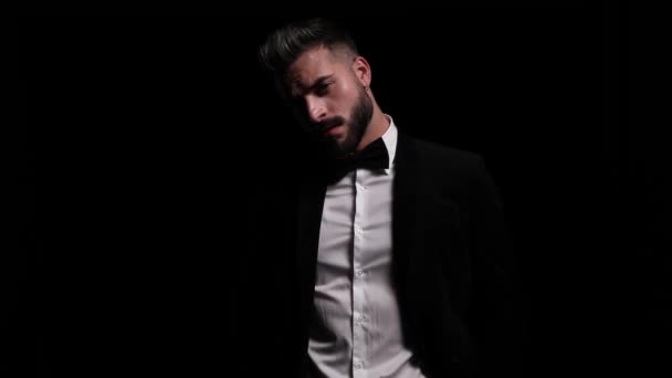 mysterious young man arranging bowtie and black tuxedo, moving in a side view position and posing, smiling, snapping fingers and leaving on black background in studio - Footage, Video