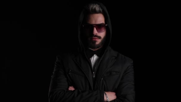 dramatic young guy in black jacket wearing sunglasses and crossing arms and looking to side on black background in studio - Footage, Video