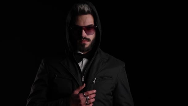 coole jongeman met een zonnebril, het regelen van zwarte jas, het vasthouden van handen in zakken en poseren op zwarte achtergrond in de studio - Video