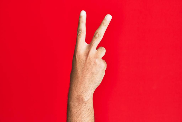 Kafkasyalı beyaz adamın kırmızı arka planda kalan kolları iki parmağını gösteriyor, işaret ediyor zafer ve kazanan sembol.  - Fotoğraf, Görsel