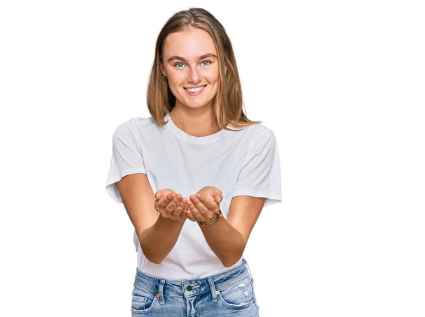 Hermosa joven rubia vistiendo camiseta blanca casual sonriendo con las palmas de las manos juntas recibiendo o dando gesto. retención y protección  - Foto, Imagen