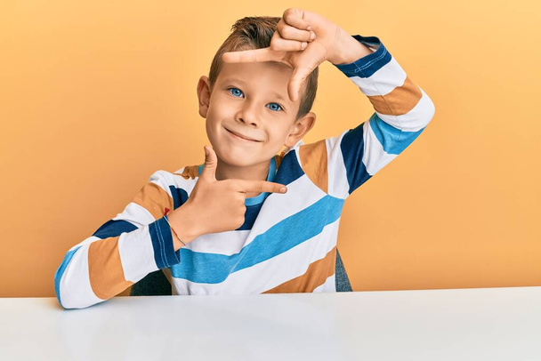 Roztomilé bělošské dítě, ležérní, sedící na stole a usmívající se a tvořící rámeček s rukama a prsty se šťastnou tváří. koncepce kreativity a fotografie.  - Fotografie, Obrázek
