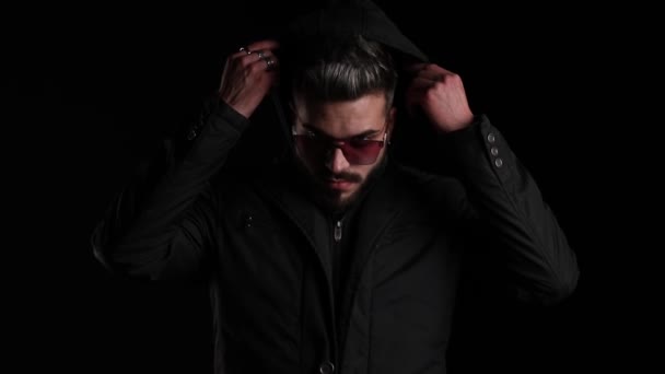 mysterieuze jongeman trekken naar beneden hoodie, vaststelling van haar, zelfverzekerd aanpassen zwarte jas en poseren op zwarte achtergrond in de studio - Video