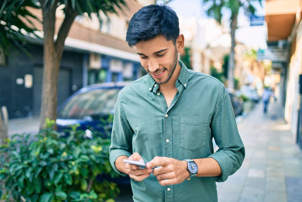 Giovane uomo latino sorridente felice utilizzando smartphone a piedi in città. - Foto, immagini