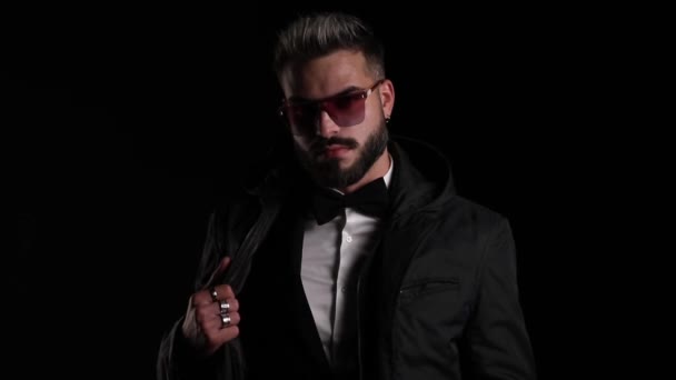sexy young guy wearing sunglasses, unzipping jacket, holding hands in pockets and looking to side, moving in a side view position and posing on black background in studio - Footage, Video