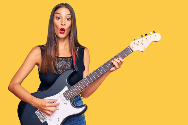 Młoda latynoska kobieta gra na gitarze elektrycznej przestraszony i zdumiony z otwartymi ustami dla zaskoczenia, niedowierzanie twarz  - Zdjęcie, obraz