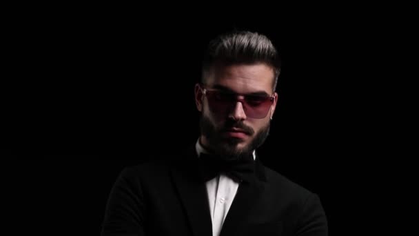zelfverzekerde elegante man met een zonnebril en zwarte smoking, armen oversteken en opzij kijken, vingers in de mond houden en stil gebaar maken op zwarte achtergrond in de studio - Video