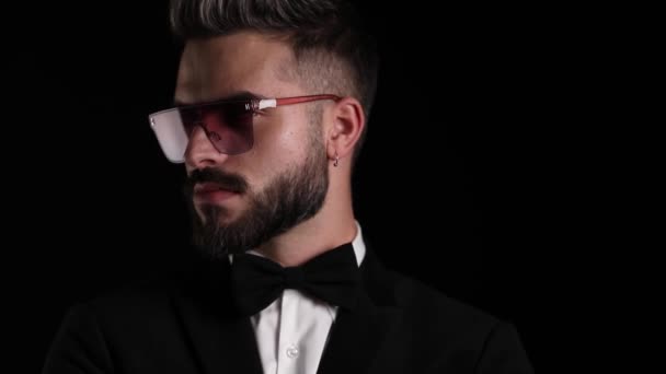 aantrekkelijke jongeman in zwarte smoking draagt een zonnebril, kijkt naar opzij, houdt sensueel de vingers aan de lippen en maakt een stil gebaar op zwarte achtergrond in de studio - Video
