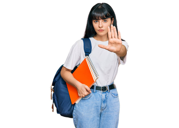 Young hispanic girl wearing student backpack and holding book with open hand doing stop sign with serious and confident expression, defense gesture  - Foto, Imagem