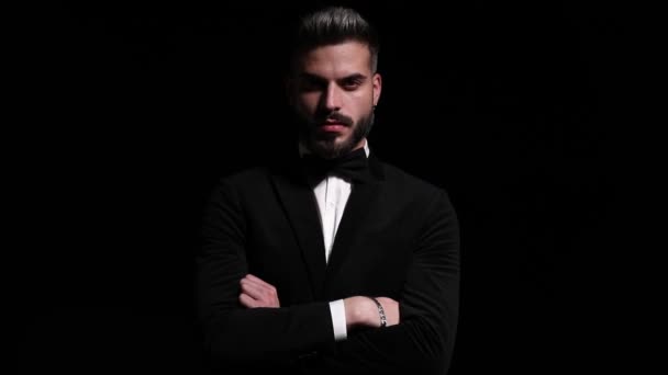 serious elegant man in black tuxedo crossing arms and looking to side, holding fingers to mouth and making silence gesture on black background in studio - Footage, Video