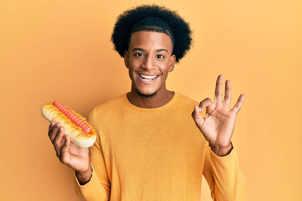 Африканский американец с африканскими волосами, поедающий хот-дог делает хорошо знак с пальцами, улыбаясь дружелюбный жест отличный символ  - Фото, изображение