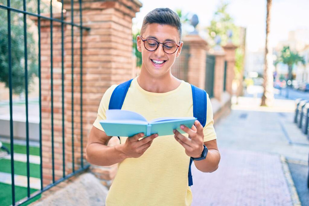 Genç İspanyol öğrenci gülümsüyor. Mutlu bir kitap okuyor. Şehrin caddesinde duruyor. - Fotoğraf, Görsel