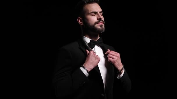 confident young man adjusting black tuxedo, crossing arms and looking to side, posing on black background in studio - Footage, Video
