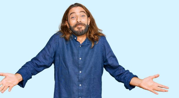 Attractive man with long hair and beard wearing casual clothes clueless and confused expression with arms and hands raised. doubt concept.  - Photo, Image