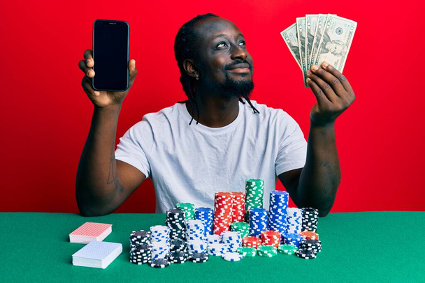 Schöner junger schwarzer Mann, der Poker spielt, Smartphone und Dollars in der Hand hält, zur Seite schaut und nachdenklich wegstarrt.  - Foto, Bild