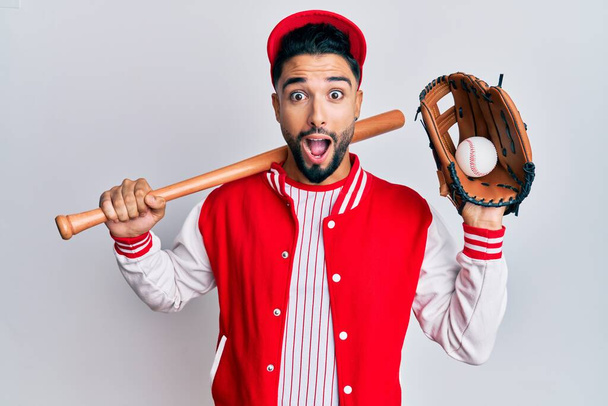 Młody człowiek z brodą gra w baseball trzymając kij i piłkę przerażony i zszokowany zaskakującą i zdumioną ekspresją, strachem i podnieconą twarzą.  - Zdjęcie, obraz