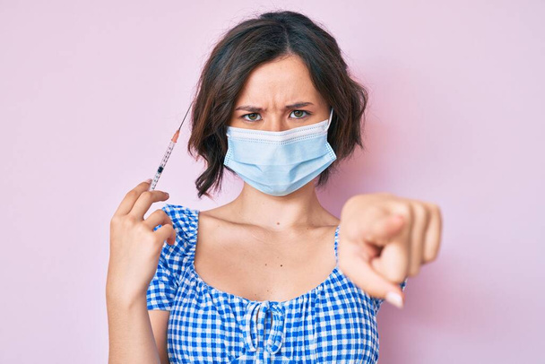 Молодая красивая женщина в медицинской маске держит шприц указывая пальцем в камеру и для вас, уверенный жест выглядит серьезным  - Фото, изображение
