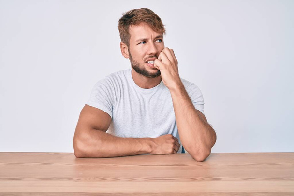 Νεαρός καυκάσιος άντρας που φοράει καθημερινά ρούχα κάθεται στο τραπέζι δείχνοντας αγχωμένος και νευρικός με τα χέρια στο στόμα να δαγκώνουν νύχια. πρόβλημα άγχους.  - Φωτογραφία, εικόνα