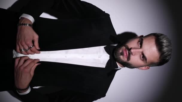 verticaal portret van een elegante jongeman die zwart fluwelen smoking aanpast, handen in zakken houdt, naar opzij kijkt en glimlacht, vingers knipt en wegloopt op een grijze achtergrond in de studio - Video