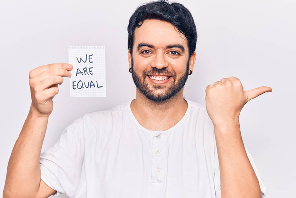 Young hispanic man holding we are equal paper pointing thumb up to the side smiling happy with open mouth  - Photo, Image