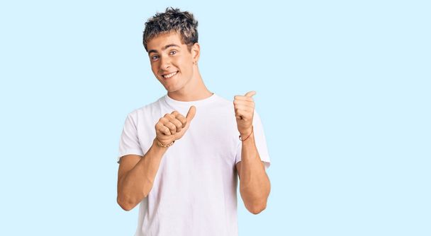 Молодой красивый мужчина в обычной белой футболке указывает на спину с поднятой рукой и большими пальцами, уверенно улыбаясь  - Фото, изображение