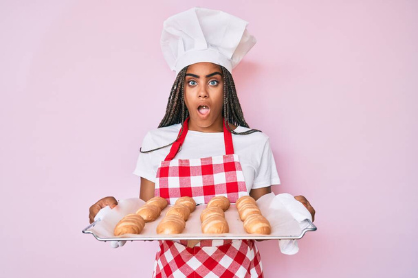 Νεαρή Αφροαμερικανή με κοτσίδες που φοράει στολή φούρναρη κρατώντας σπιτικό ψωμί στο πρόσωπο, δείχνοντας σκεπτική και σαρκαστική, έκπληκτη με ανοιχτό στόμα  - Φωτογραφία, εικόνα
