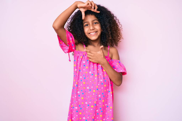 Afro-Amerikaanse kind met krullend haar dragen casual jurk glimlachend maken frame met handen en vingers met gelukkig gezicht. creativiteit en fotografie.  - Foto, afbeelding