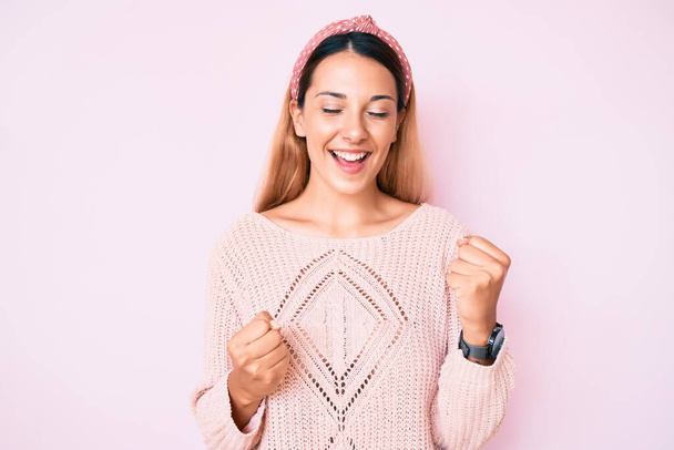 Молодая брюнетка в повседневной одежде на розовом фоне празднует удивление и удивление за успех с поднятыми руками и закрытыми глазами  - Фото, изображение