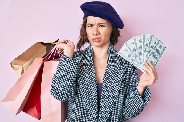 Mujer hermosa joven con boina sosteniendo bolsas de compras y billetes de dólares expresión despistada y confusa. concepto de duda.  - Foto, imagen