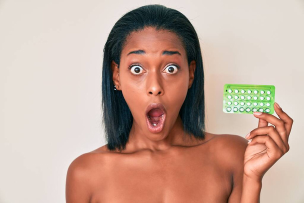 Młoda Afroamerykanka trzymająca tabletki antykoncepcyjne robi rybi pysk ustami i mrużącymi oczami, szalona i komiczna.  - Zdjęcie, obraz