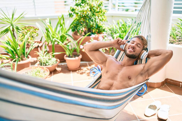 Όμορφος καυκάσιος άντρας χαμογελάει χαρούμενος αναπαύεται σε μια αιώρα στη βεράντα ακούγοντας μουσική φορώντας ακουστικά - Φωτογραφία, εικόνα