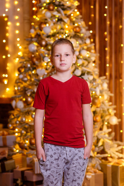An eight-year-old European boy in a burgundy T-shirt stands in the background of a Christmas tree and looks into the camera. Christmas portrait of a boy - Foto, afbeelding