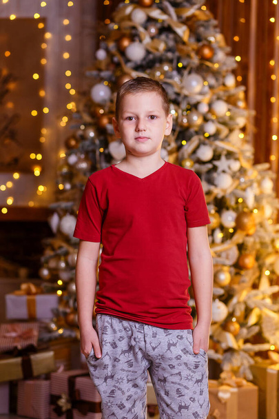An eight-year-old European boy in a burgundy T-shirt stands in the background of a Christmas tree and looks into the camera. Christmas portrait of a boy - Valokuva, kuva