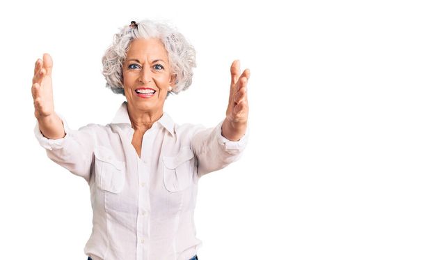 Femme âgée aux cheveux gris portant des vêtements décontractés regardant la caméra souriant à bras ouverts pour un câlin. expression joyeuse embrassant le bonheur.  - Photo, image