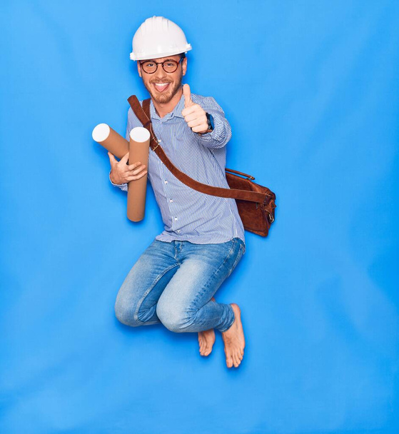 Young handsome architect man wearing leather bag and hardhat smiling happy. Jumping with smile on face holding blueprints doing ok sign with thumb up over isolated blue background. - Photo, Image