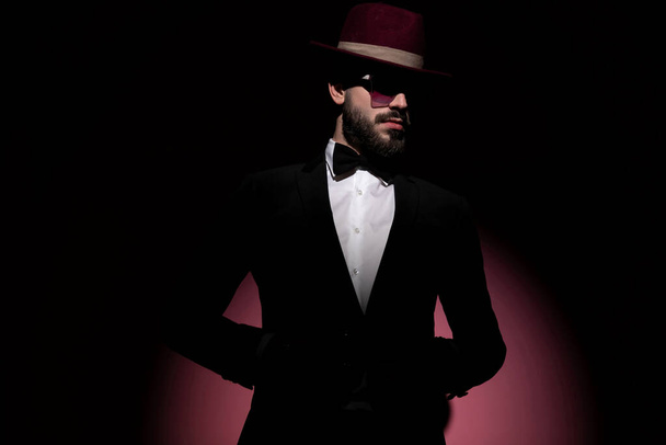 attraente uomo d'affari che chiude la giacca, distogliendo lo sguardo e indossando un cappello bordeaux - Foto, immagini