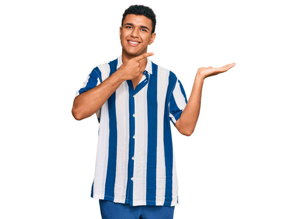 Junger arabischer Mann in lässiger Kleidung staunt und lächelt in die Kamera, während er mit der Hand präsentiert und mit dem Finger zeigt.  - Foto, Bild