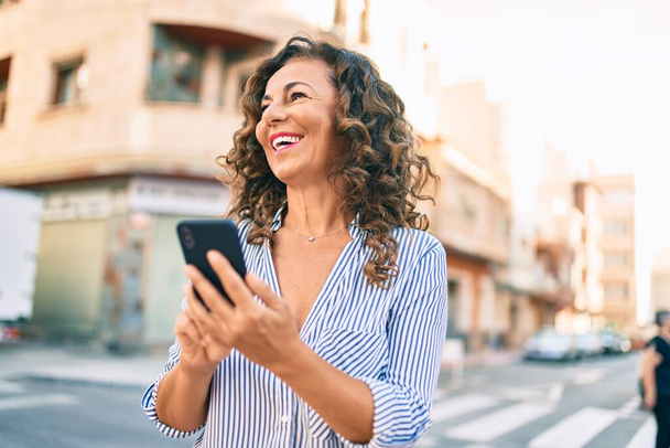 Mujer hispana de mediana edad sonriendo feliz y usando smartphone en la ciudad. - Foto, Imagen