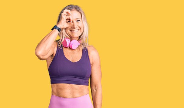 Middle age fit blonde woman wearing gym clothes and using headphones doing ok gesture with hand smiling, eye looking through fingers with happy face.  - Фото, зображення