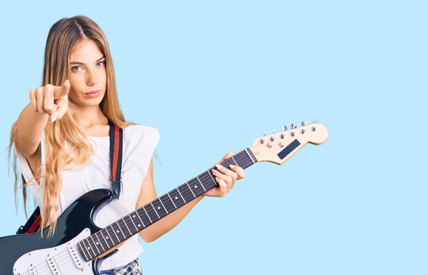 Beautiful caucasian woman with blonde hair playing electric guitar pointing with finger to the camera and to you, confident gesture looking serious  - Φωτογραφία, εικόνα