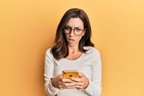 Jonge brunette vrouw met behulp van smartphone over gele achtergrond in shock gezicht, op zoek sceptisch en sarcastisch, verrast met open mond  - Foto, afbeelding