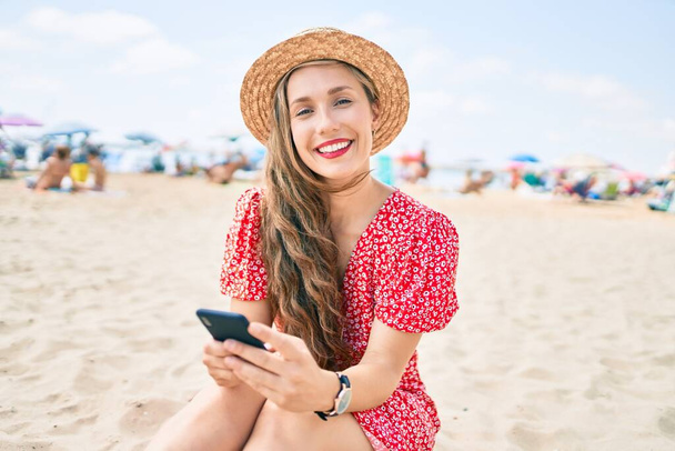 Молодая блондинка в отпуске с помощью смартфона сидит на песке на пляже - Фото, изображение